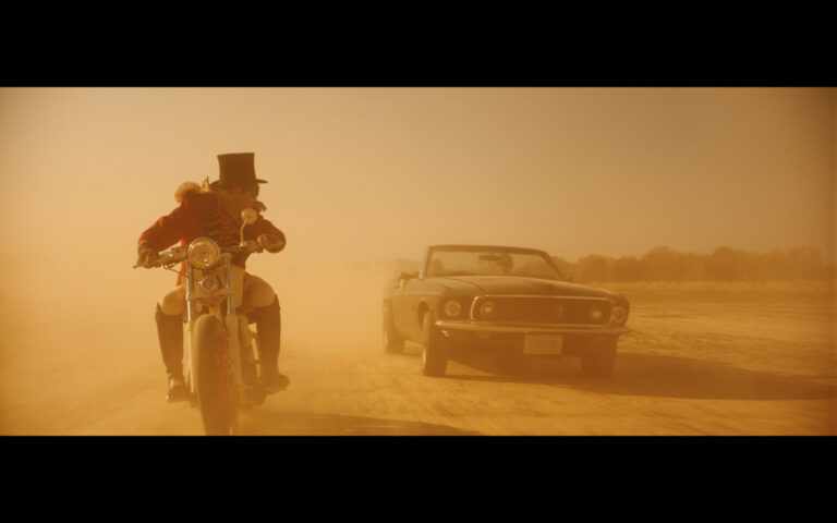 Man on motorcycle and black Mustang in dusty desert.