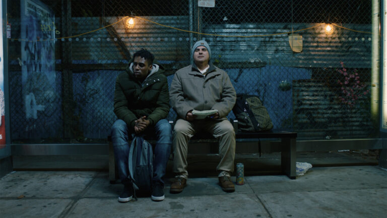 Two men sit on a bench at night, one holding cash.