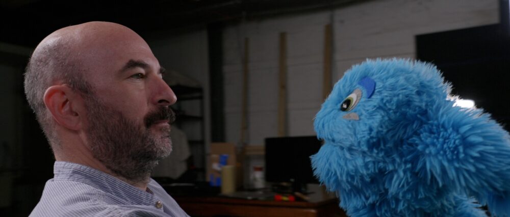 Man with beard looking at a blue puppet. I hope this helps!