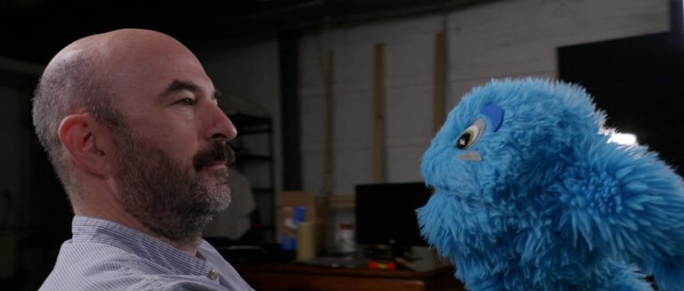 Man with beard looking at a blue puppet. I hope this helps!