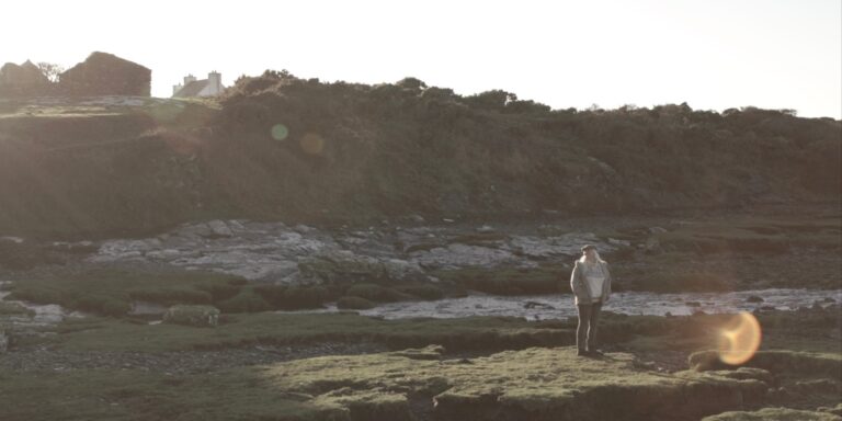 Woman stands in a coastal landscape near old stone buildings, possibly related to "The Beekeeper" blog.