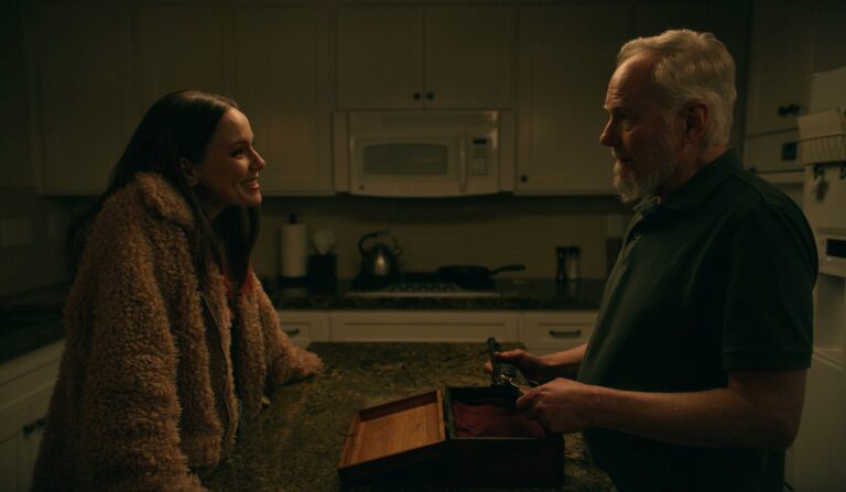 Woman in fuzzy coat smiles at man with wooden box in kitchen.