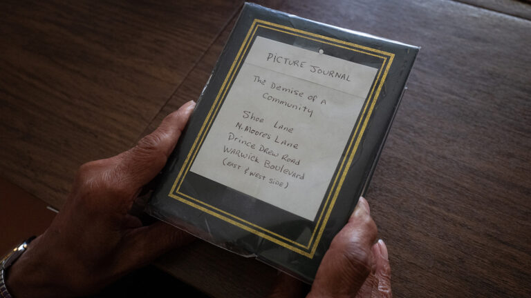 Hands holding a "Picture Journal" titled "The Demise of A Community," listing streets: Shoe Lane, N. Moores Lane, etc.