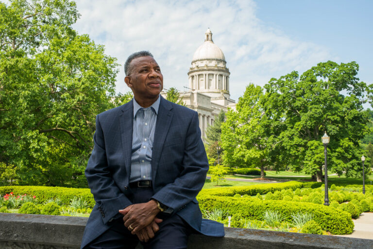 Black man in a blue suit sits in front of the Kentucky State Capitol.