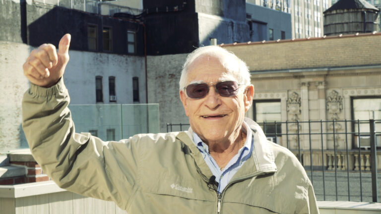 Elderly man gives a thumbs up on a rooftop in a city setting.