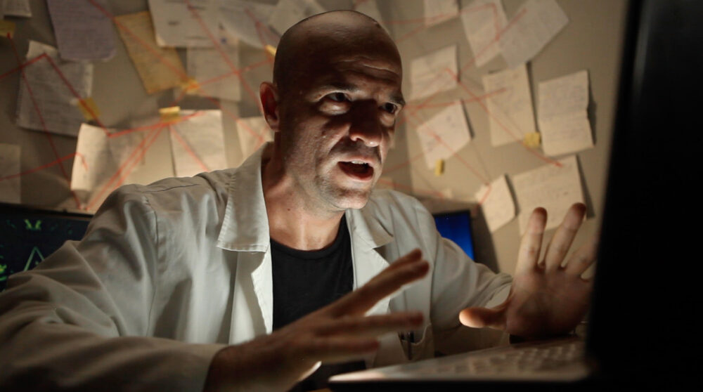 Frustrated man in lab coat stares at laptop with conspiracy wall in background.