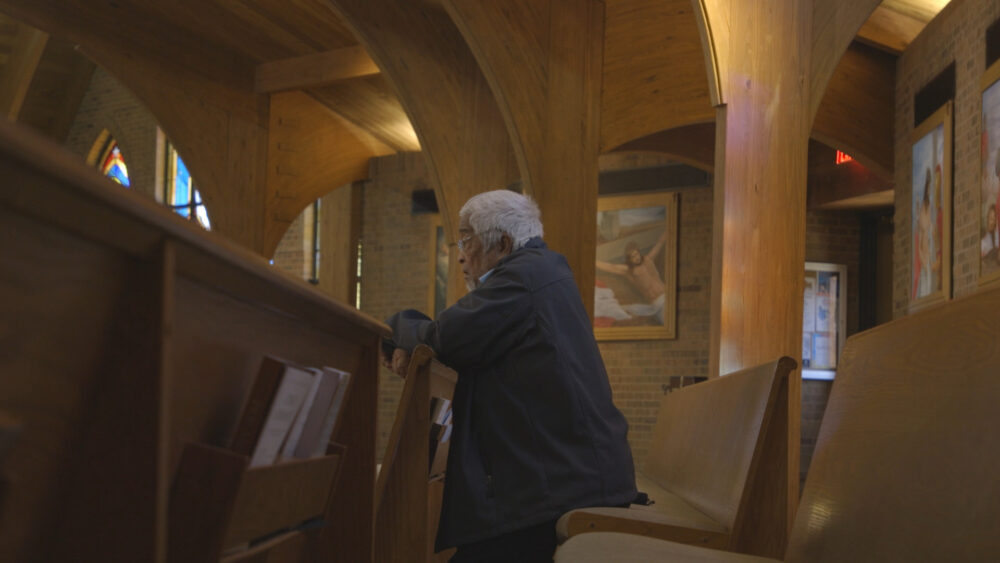 Man prays in church pew, with religious art in background.