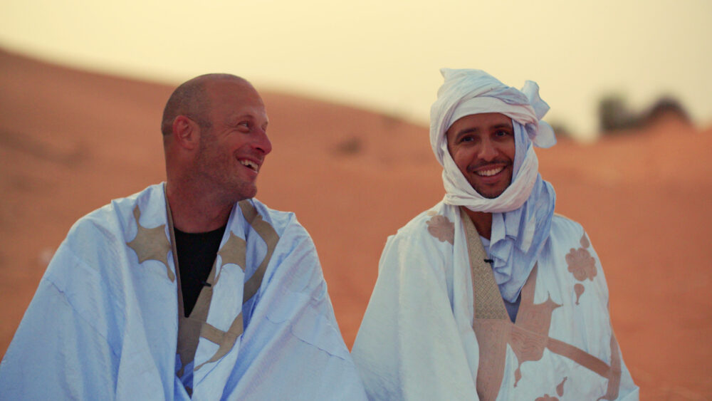 Two men in traditional Berber clothing smile in the Sahara desert.