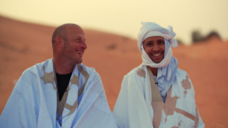Two men in traditional Berber clothing smile in the Sahara desert.