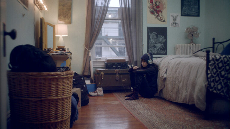 Teen with headphones sits on floor in a cluttered, vintage bedroom.