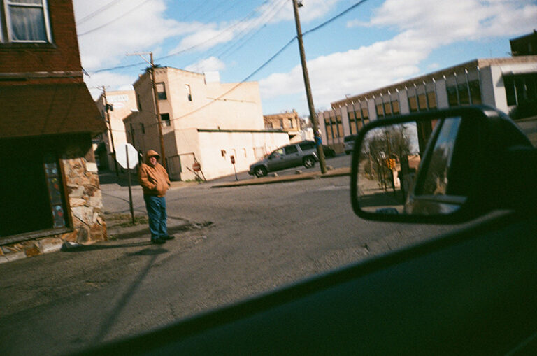 Man stands on a street corner in a small town, seen from a car window. "Still Home" evokes a sense of place.