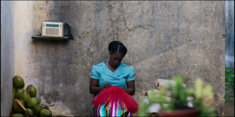 Woman sewing red fabric outdoors, radio on shelf, coconuts nearby.