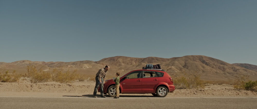 Father and son by a red car on a desert road, with luggage on top.