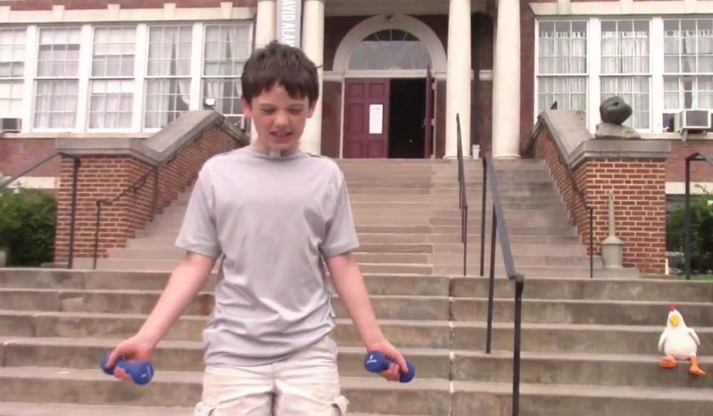 Boy exercising with dumbbells outside McGuffey Art Center, with a chicken plush toy sitting on the stairs nearby.
