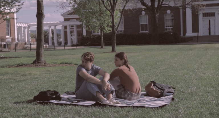 Young couple sitting on a blanket in a grassy campus setting, enjoying a moment together.