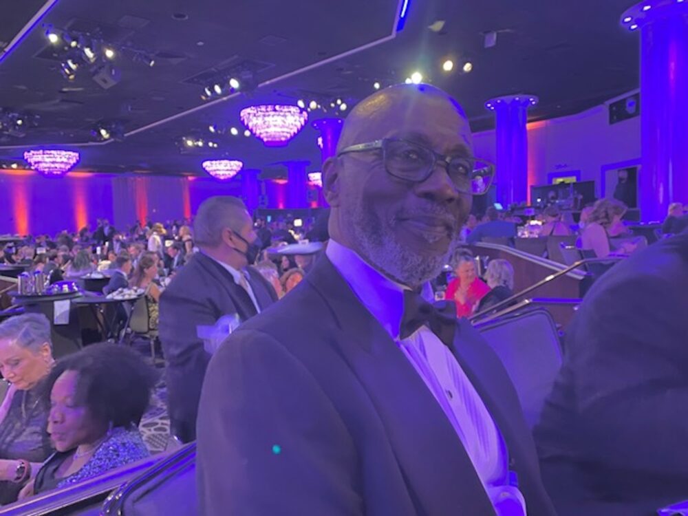 Preston Holmes at an event, wearing a tuxedo and glasses, surrounded by a crowd.