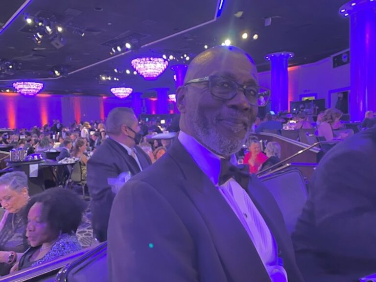 Preston Holmes at an event, wearing a tuxedo and glasses, surrounded by a crowd.