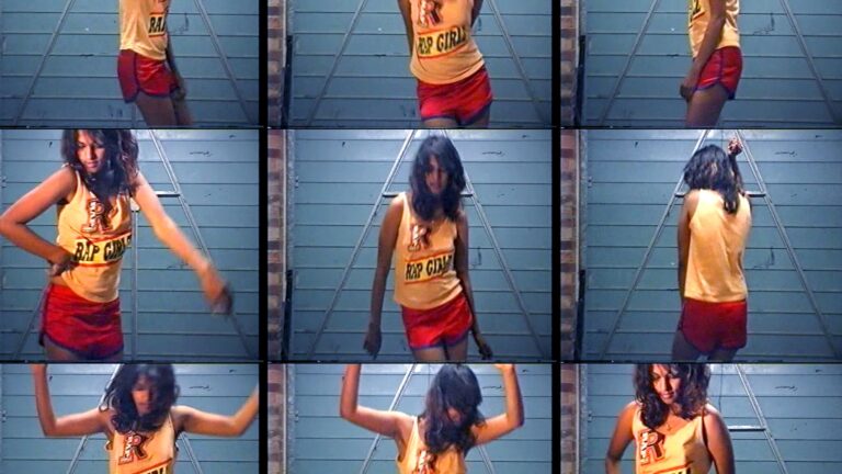 M.I.A. wearing a "Rap Girl" tank top and red shorts dancing in front of a blue garage door.