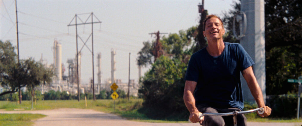 Man on bike, possibly from "Red Rocket," smiles near industrial site.