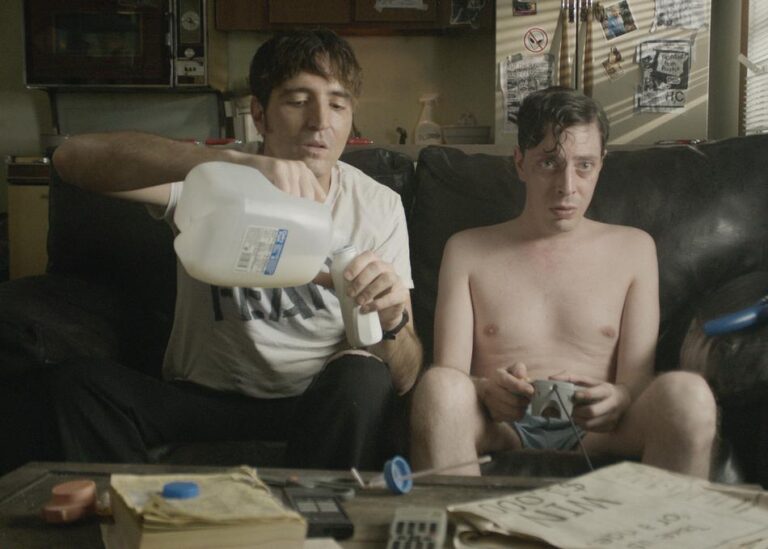 Man pouring milk for another man playing video games on a couch.