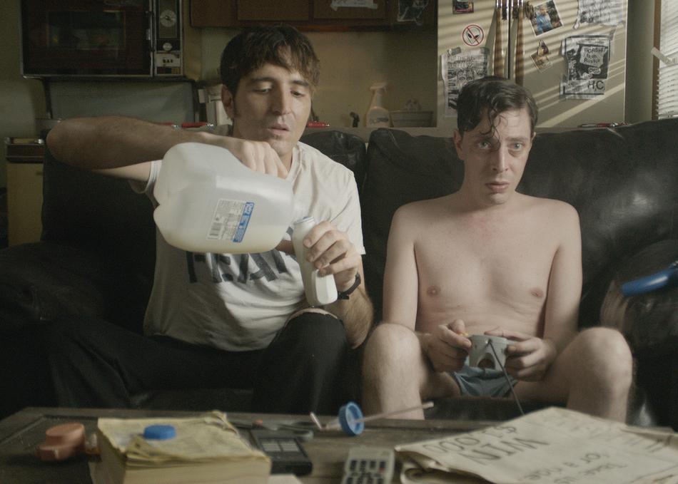 Man pouring milk for another man playing video games on a couch.