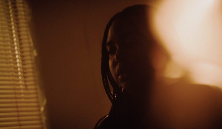 Pensive young Black woman in dark, warm lighting with window blinds.