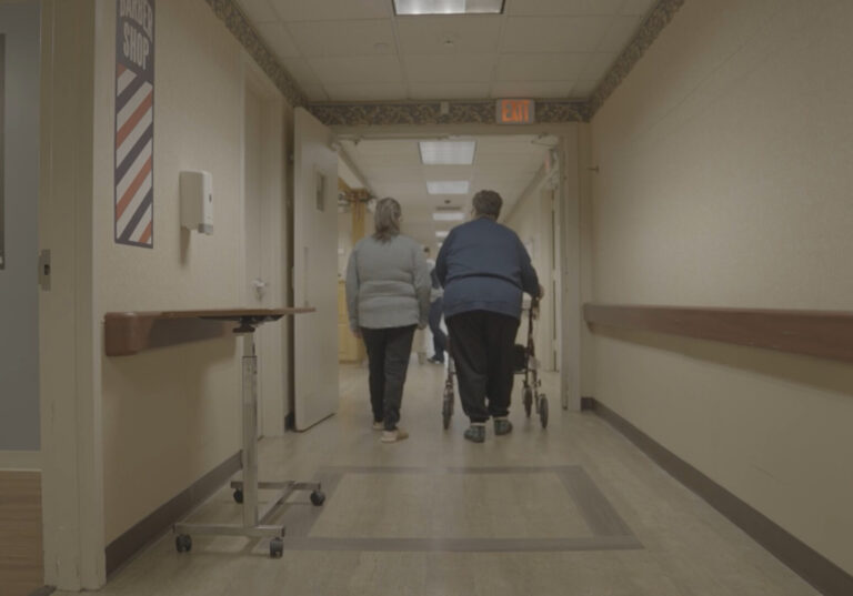 Two people walking down a nursing home hallway with a walker.