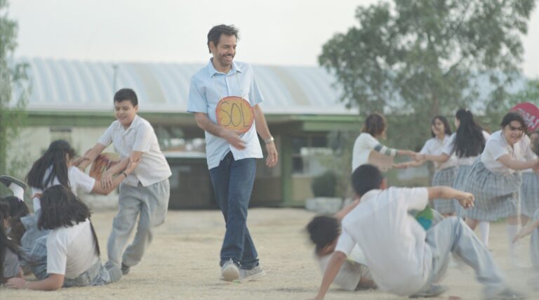 Teacher smiles during a schoolyard game with students. Sign reads "SOL.