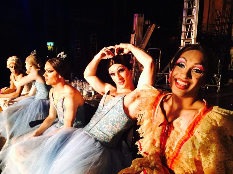 Drag ballet dancers in costumes backstage, smiling for the camera.
