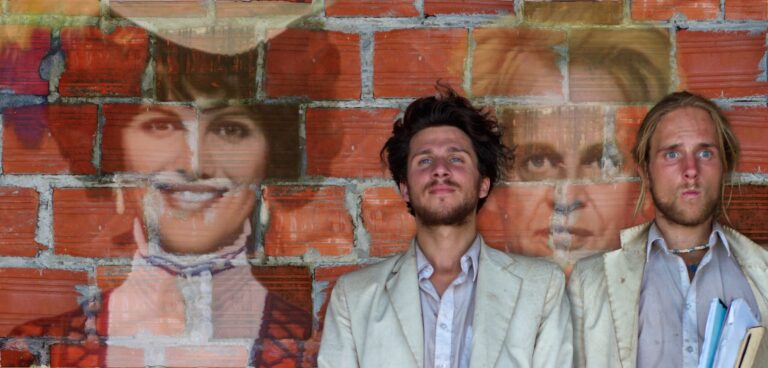 Two men in suits stand before a brick wall with faded portrait artwork behind them.