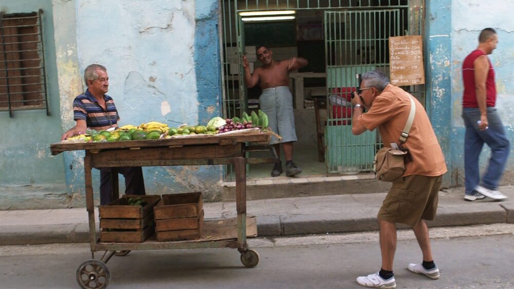 Cuban street scene: Man photographs fruit vendor near a shop, evoking Walker Evans' style in "Return to Cuba"