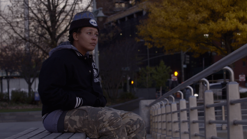 Young person sits on a bench in a park, wearing a Brooklyn jacket and hat.