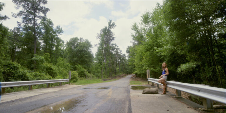 Woman on roadside bridge with bag. Rural road and trees.