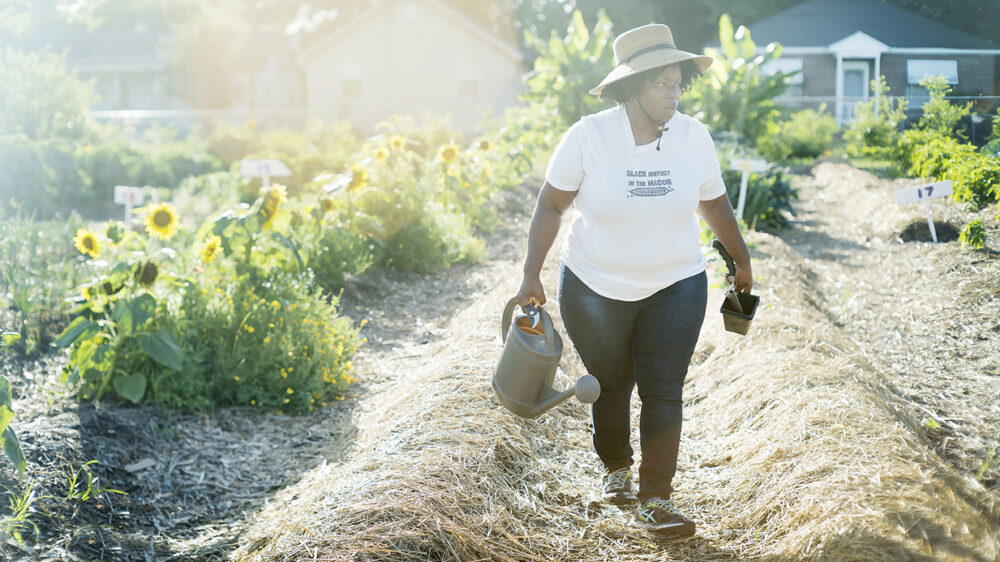 Woman gardening with watering can and plants in a community garden. Shirt reads "Black Whitney in the Garden".