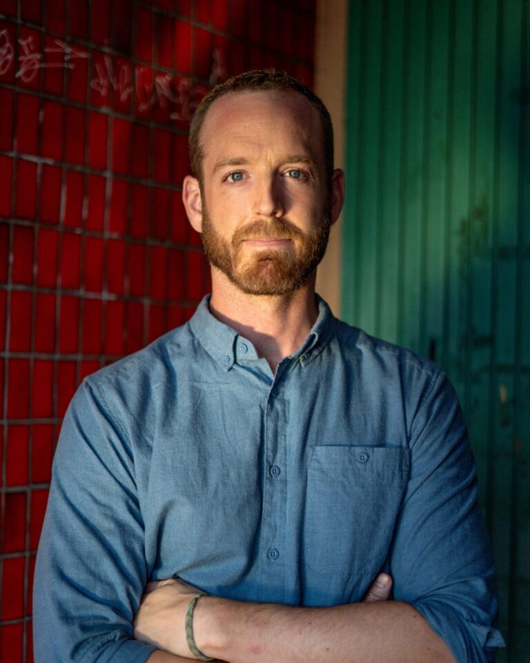 Portrait of Scott Faris with arms crossed, standing in front of a red and green wall.