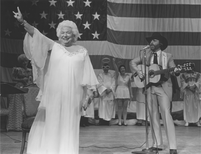 Liz Carpenter on stage with a musician and American flag backdrop, performing.