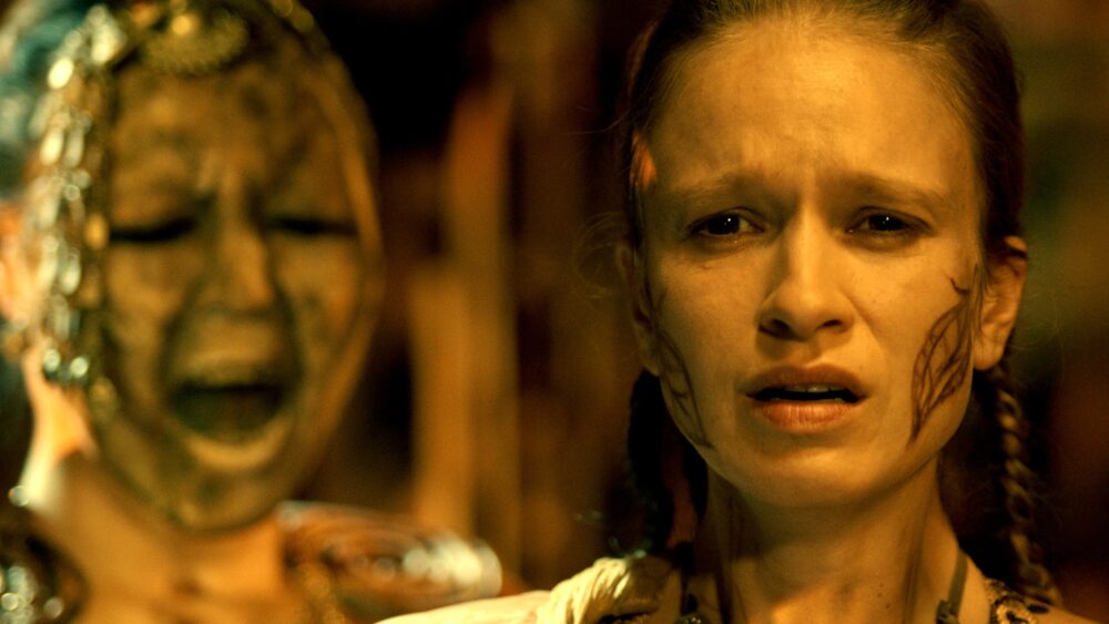 Distressed woman with face paint, background figure screaming.