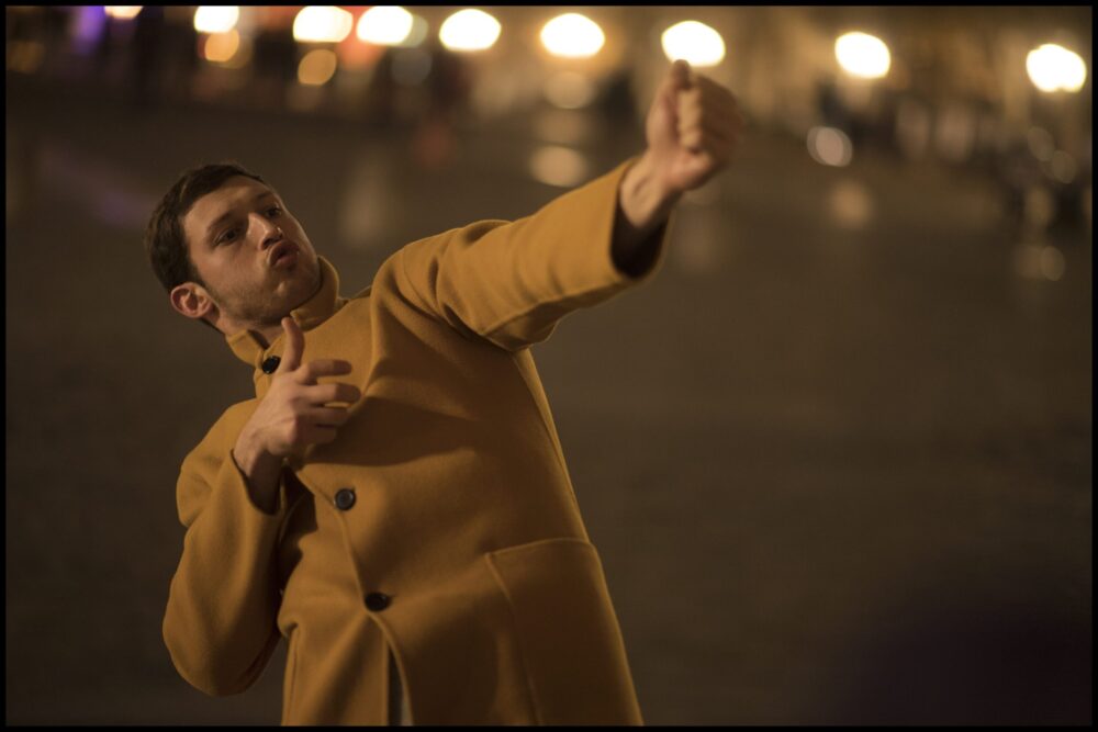 Man in a mustard coat gesturing outdoors at night.