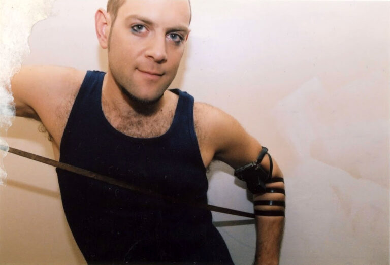 Man in tank top with arm bands and dark eye makeup.
