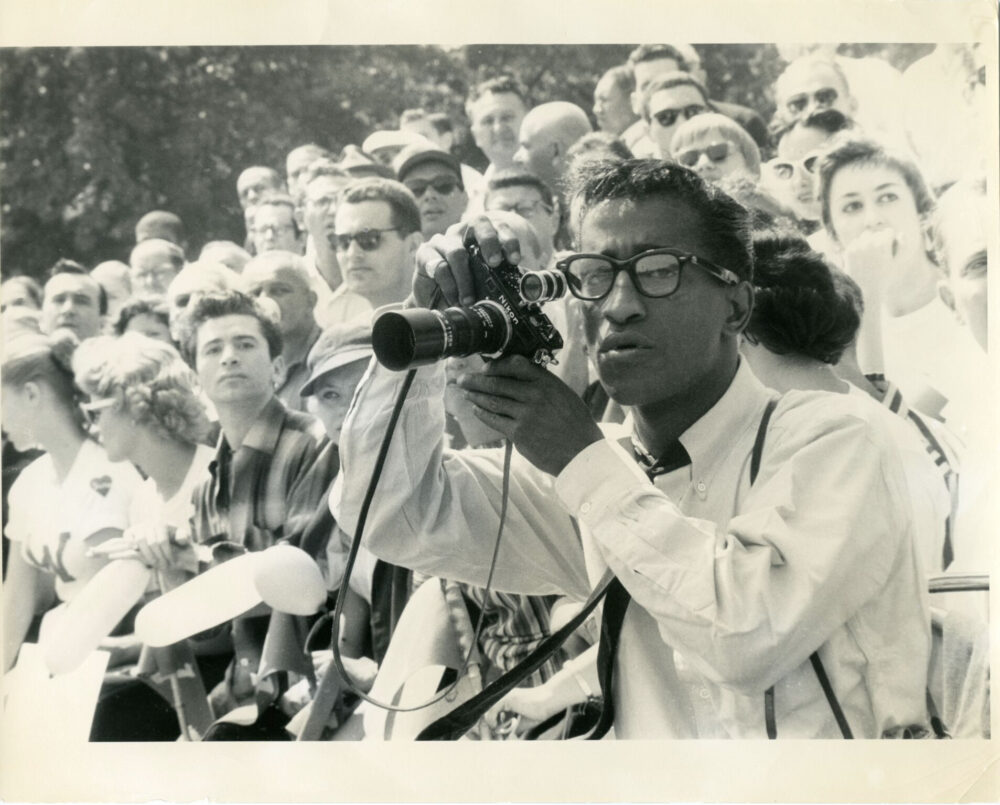 Sammy Davis Jr. taking a photo with a Nikon camera in a crowd.