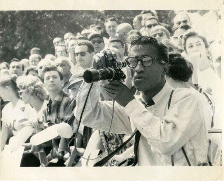 Sammy Davis Jr. taking a photo with a Nikon camera in a crowd.