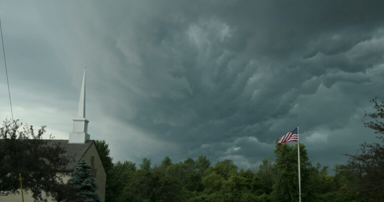 Church steeple and American flag under a stormy sky.
