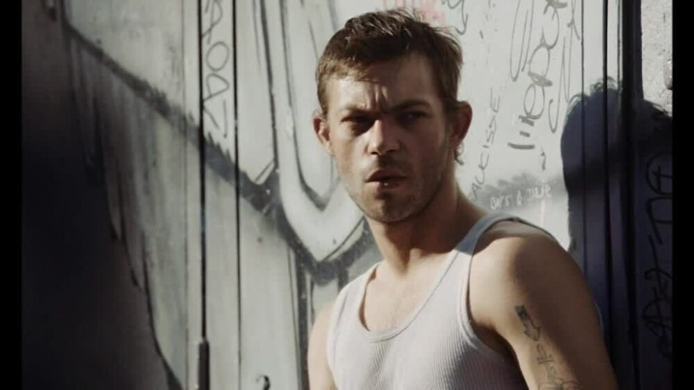 Man in white tank top stands against graffiti wall.