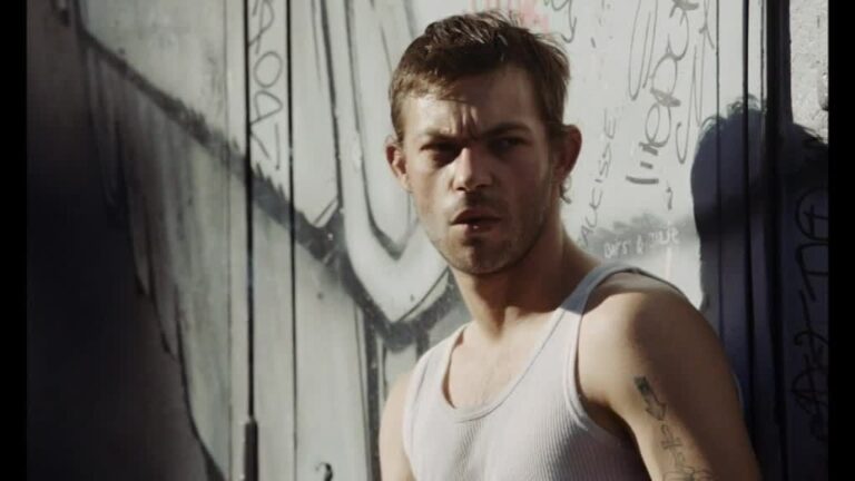 Man in white tank top stands against graffiti wall.