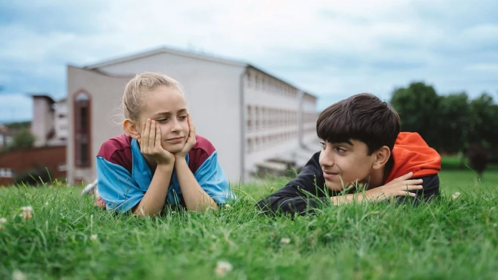 Two young friends lying on a grassy hill, looking at each other.