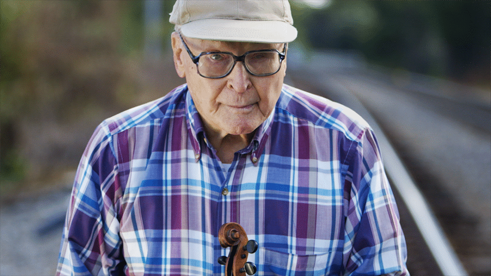 Elderly man with violin near train tracks, wearing a cap and plaid shirt.