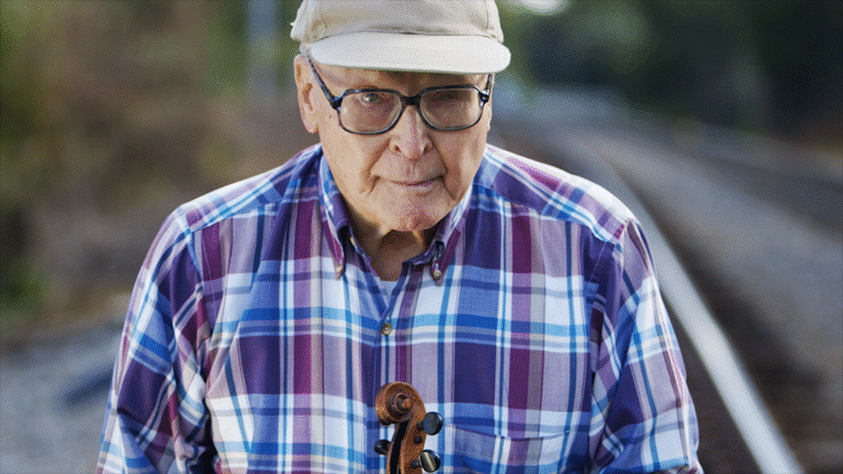 Elderly man with violin near train tracks, wearing a cap and plaid shirt.