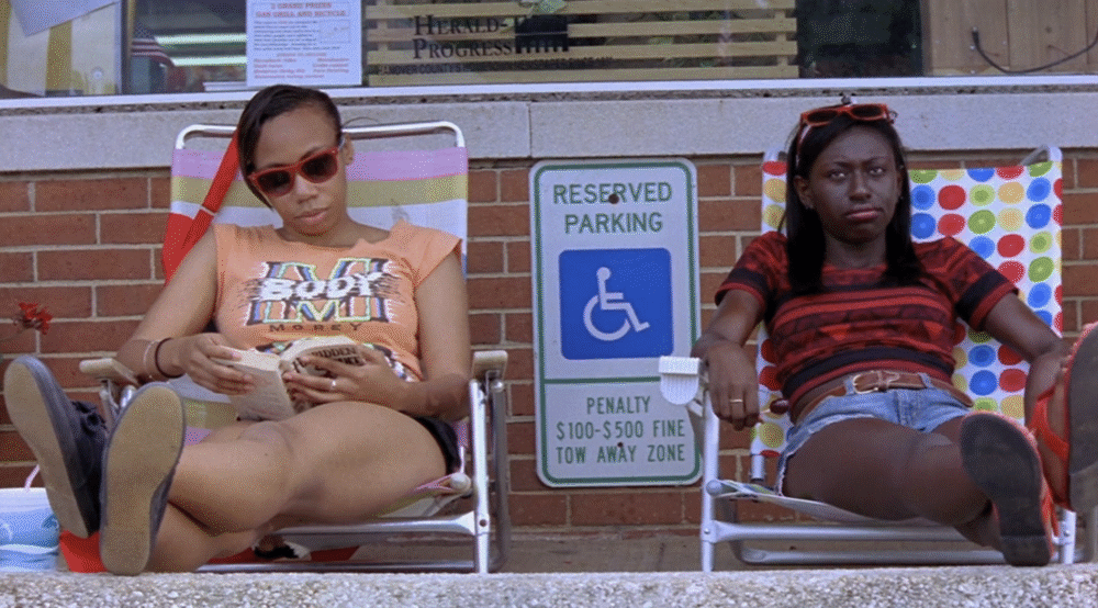 Two women relax in chairs near a "Reserved Parking" sign, reading a newspaper.