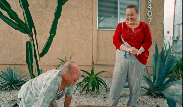Comedic scene: Man sticking tongue out at woman in paint-splattered overalls outside building.