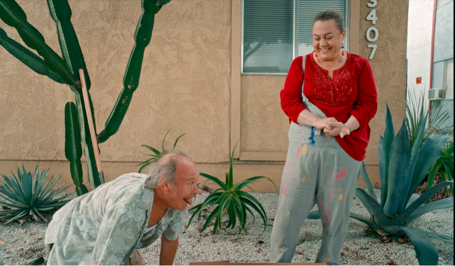 Comedic scene: Man sticking tongue out at woman in paint-splattered overalls outside building.